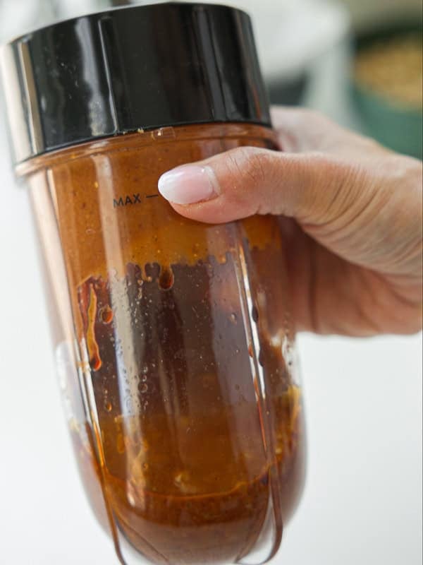 A hand holding a blender cup filled with reddish-brown sauce, showing the liquid level just below the MAX line marked on the cup.